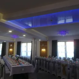 Sala bankietowa z długimi stołami, krzesła z dekoracyjnymi pokrowcami, oświetlenie sufitowe LED w kolorze niebieskim i żyrandole.