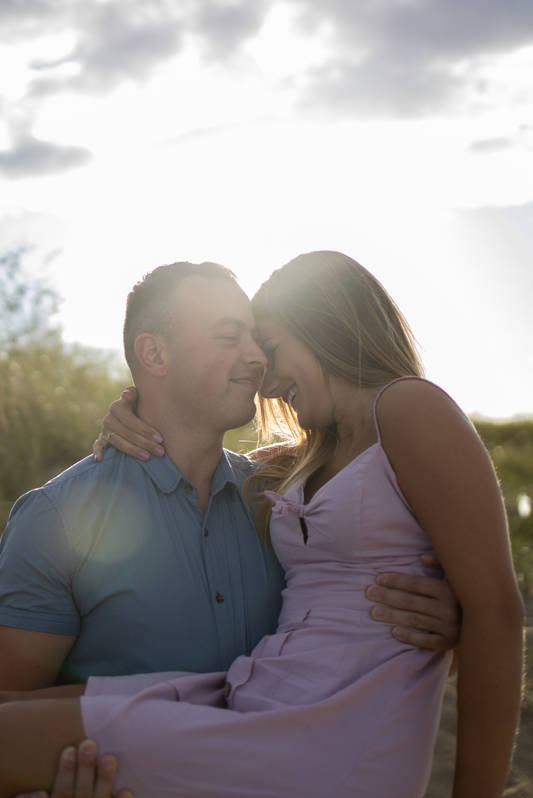 Uśmiechnięta kobieta w jasnoróżowej sukience niesiona przez mężczyznę na tle rozświetlonego słońcem krajobrazu.