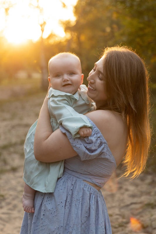 Uśmiechnięta matka trzyma niemowlę w pastelowych ubraniach na tle zachodzącego słońca i drzew.
