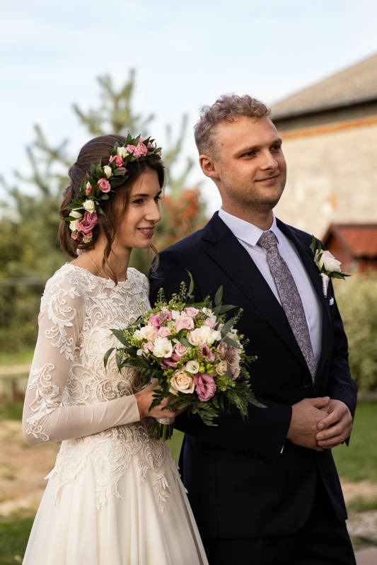 Para młoda: Panna młoda w koronkowej sukni i wianku z róż, trzyma bukiet, obok pan młody w granatowym garniturze i wzorzystym krawacie.