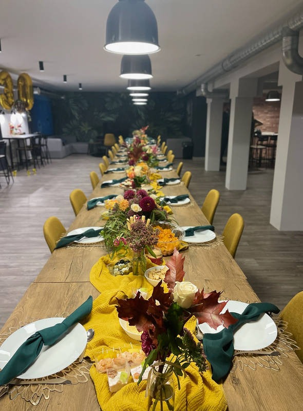 Długi stół z drewna, nakryty na uroczystość, z dekoracjami z kwiatów, liści i żółtej tkaniny, zielone serwetki na białych talerzach, oświetlenie sufitowe, balony z cyframi w tle.
