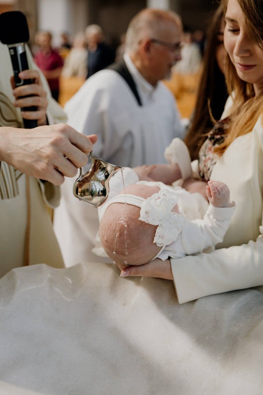 Ksiądz polewa wodą głowę niemowlęcia trzymanego przez kobietę podczas ceremonii chrztu, widoczny mikrofon w dłoni kapłana.
