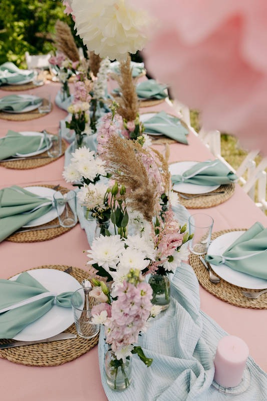 Dekoracja stołu na przyjęciu: różowy obrus, jutowe podkładki, białe talerze, miętowe serwetki, szklanki, kompozycje z białych i różowych kwiatów w małych wazonach, trawa pampasowa oraz pastelowy...