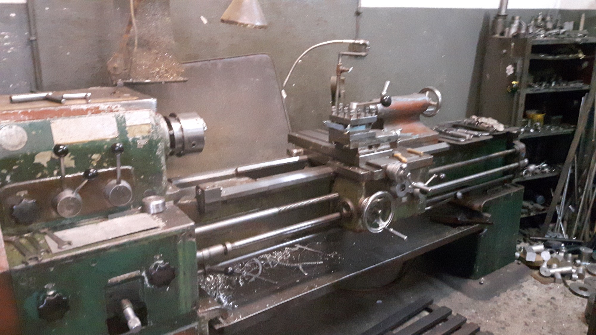 Stary, zielony tokarka w warsztacie, widoczne wióry i narzędzia na półkach w tle.