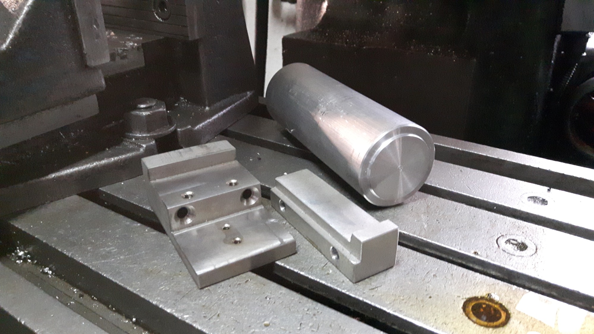 Surowe, metalowe elementy obrabiane na frezarce, w tym cylinder z widocznymi śladami obróbki i blok z otworami, leżące na stole roboczym maszyny.