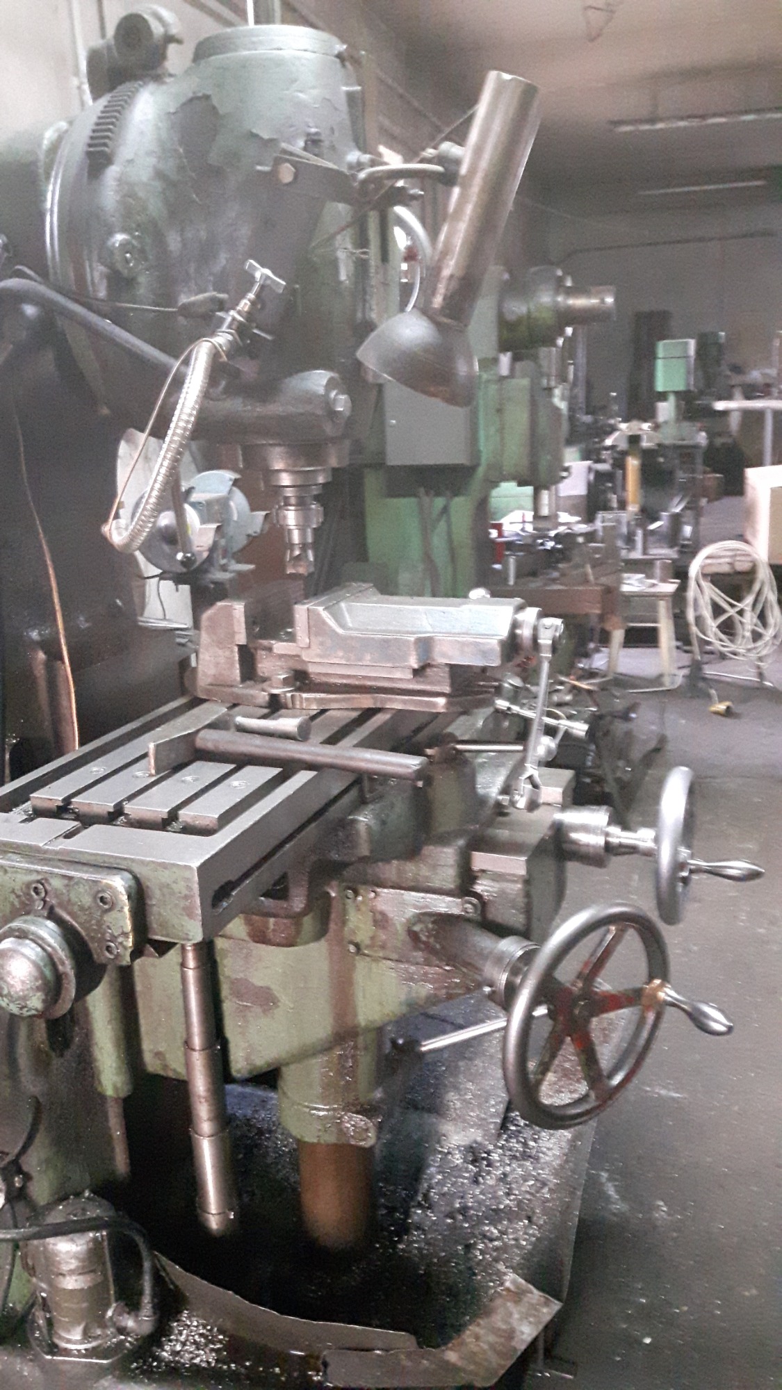Stara, zielona frezarka pionowa z zamocowanym elementem obrabianym, widoczne wióry metalowe i inne maszyny w tle.