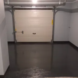 Gładka, ciemna posadzka w garażu z częściowo otwartą, segmentową bramą garażową w odcieniach beżu; widoczne ślady świeżo wylanej powłoki i wałek malarski w rogu.