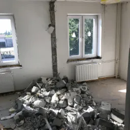 Wnętrze po częściowej rozbiórce ściany działowej, z widocznym stosem gruzu na podłodze, odsłoniętą instalacją elektryczną i żółtą lampą w tle.