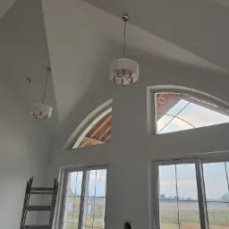 Wnętrze z wysokim sufitem po malowaniu na biało, widoczne okna łukowe, drabina i lampy. Ściany świeżo pomalowane, z widocznymi otworami na instalacje.