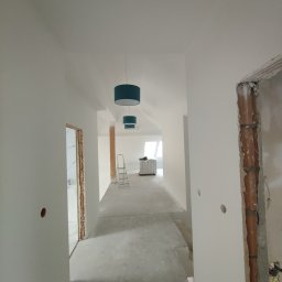 Usługi Remontowo-Budowlane JAK-BUD Jakub Balcer - Długi korytarz w jasnych barwach po remoncie, z widoczną drabiną i materiałami budowlanymi w tle. Otwory po gniazdkach w ścianach, brakujące ościeżnice.