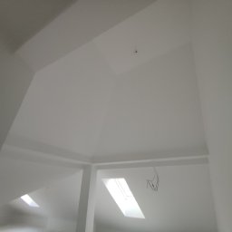 Usługi Remontowo-Budowlane JAK-BUD Jakub Balcer - Białe, świeżo pomalowane poddasze ze skosami i oknem dachowym. Widoczne przewody elektryczne i fragment ściany z nieostrym elementem. Neutralna kolorystyka.