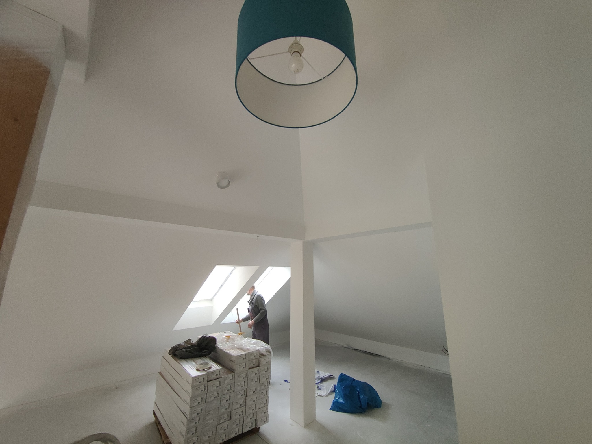 Adaptacja poddasza: malowanie na biało w skosach, widoczny pracownik z wałkiem, paleta z farbami, niebieski worek i lampa sufitowa w kolorze morskim.