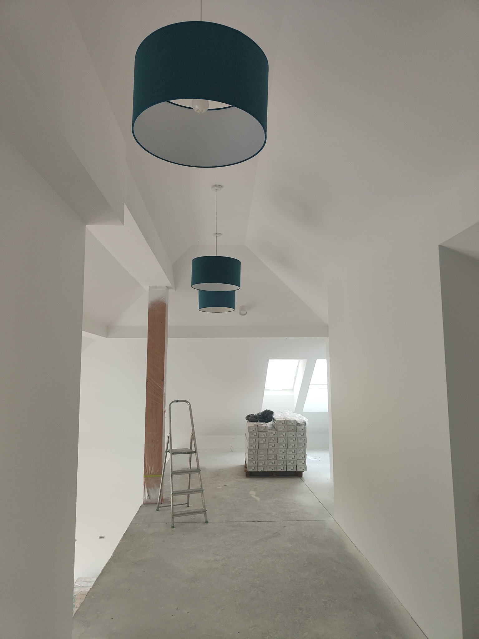 Białe, nowo pomalowane poddasze z turkusowymi lampami. Na podłodze paleta z materiałami budowlanymi, obok drabina. Widok w głąb pomieszczenia z oknem dachowym.