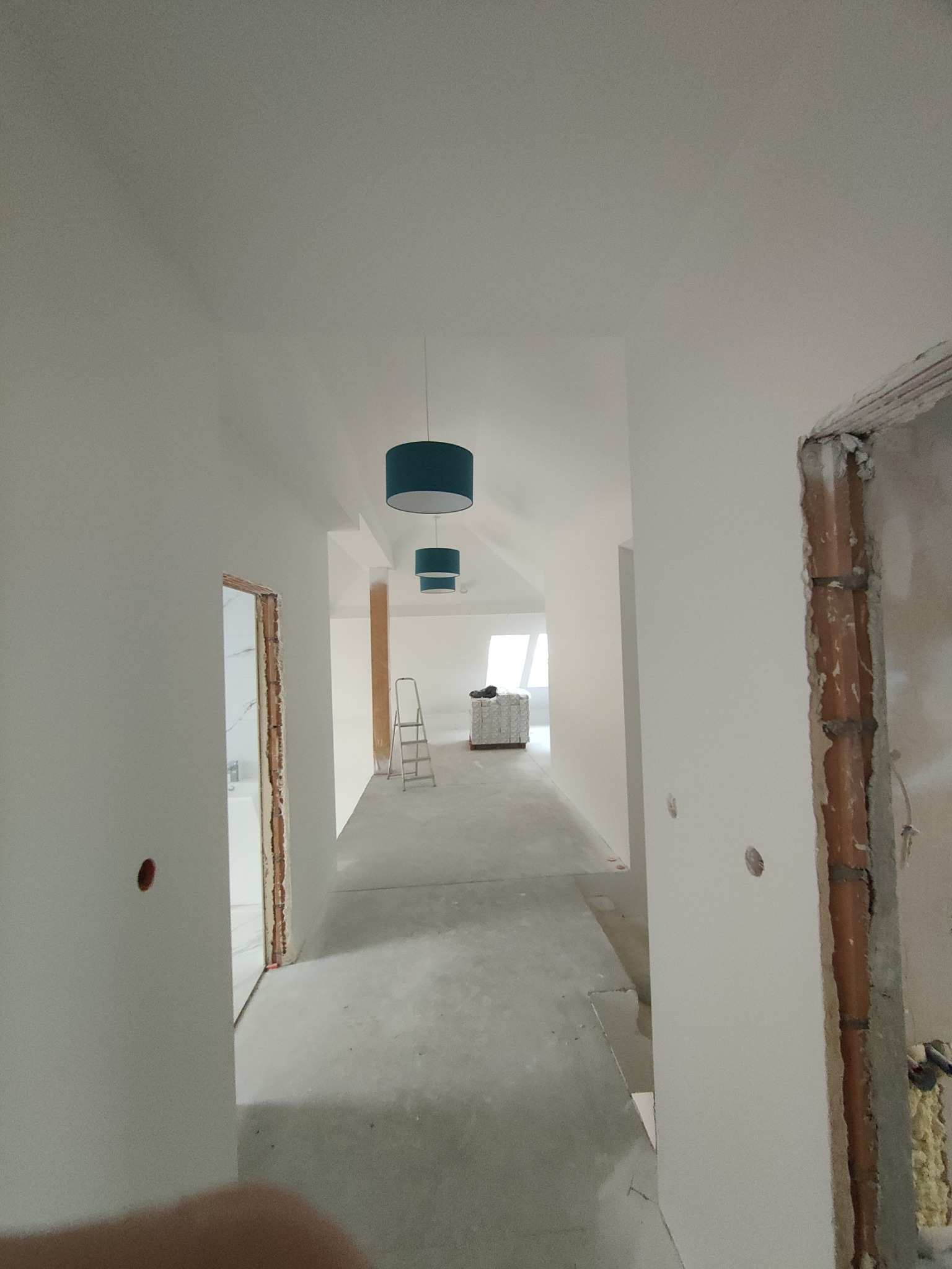 Długi korytarz w jasnych barwach po remoncie, z widoczną drabiną i materiałami budowlanymi w tle. Otwory po gniazdkach w ścianach, brakujące ościeżnice.