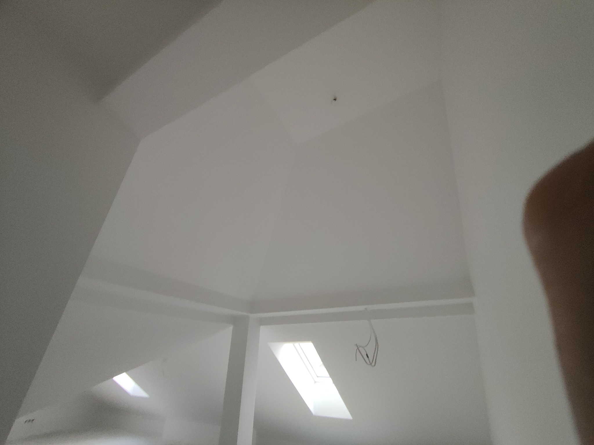 Białe, świeżo pomalowane poddasze ze skosami i oknem dachowym. Widoczne przewody elektryczne i fragment ściany z nieostrym elementem. Neutralna kolorystyka.