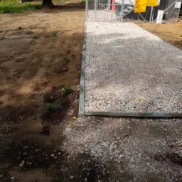 Utwardzony żwirem teren z obrzeżami z kostki betonowej prowadzący do ogrodzonego obiektu technicznego z żółtą tablicą informacyjną.