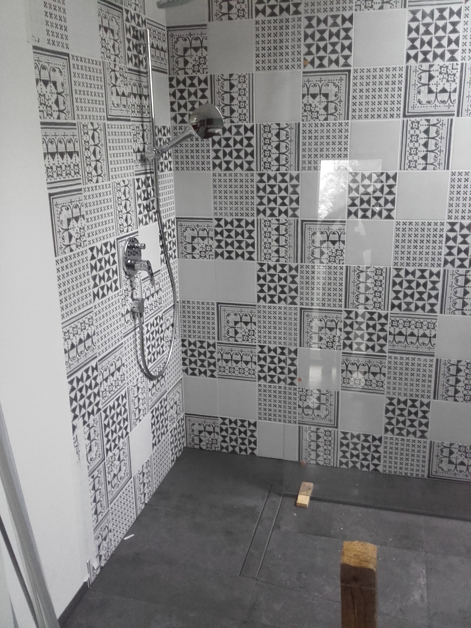 Wykończona łazienka z czarno-białymi, wzorzystymi płytkami na ścianach i szarą podłogą z odpływem liniowym, widoczna bateria prysznicowa i deszczownica, fragment ściany pomalowanej na biało.
