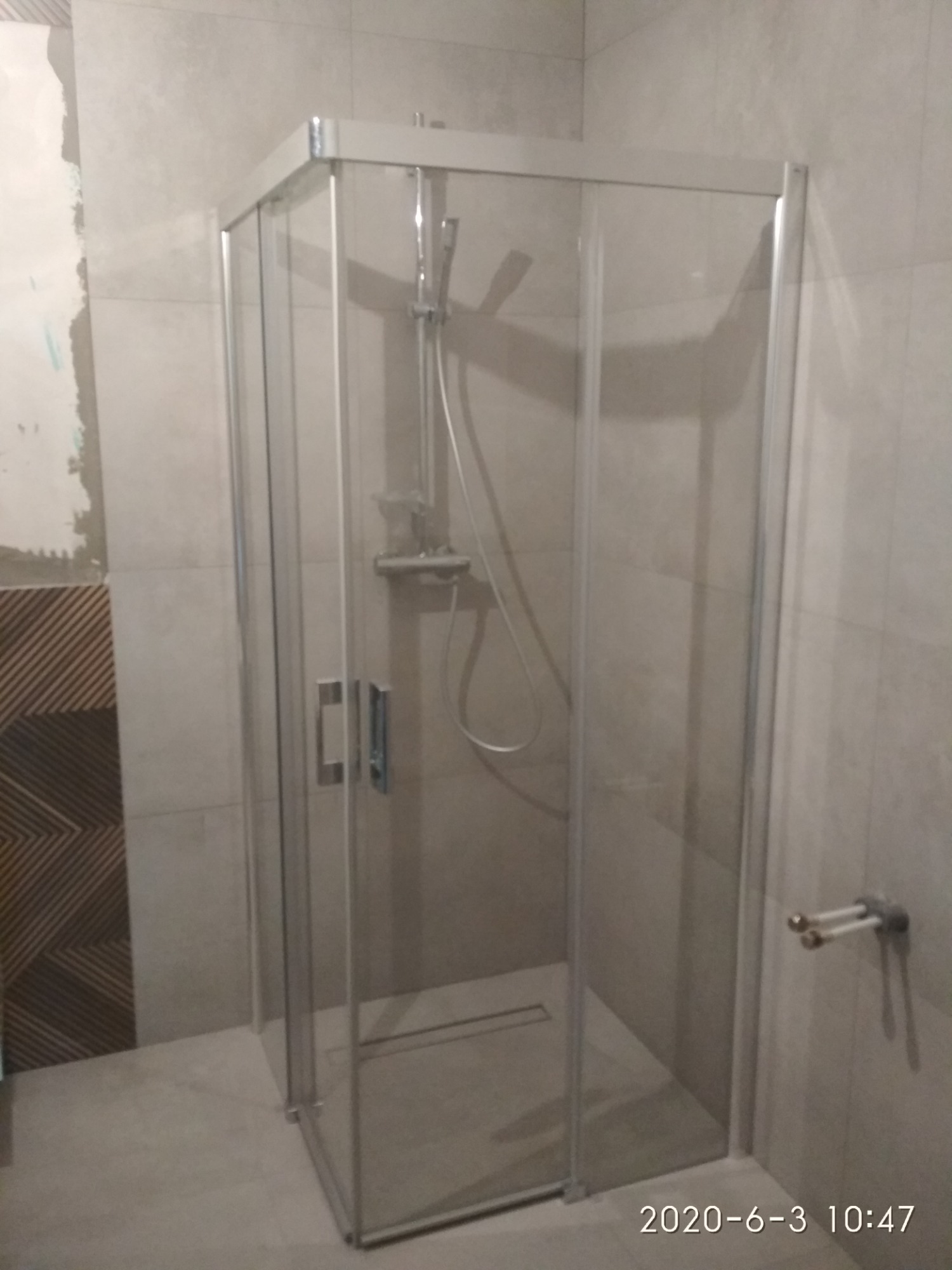 Szklana kabina prysznicowa z chromowanymi profilami w trakcie montażu, widoczne niedokończone prace glazurnicze na ścianie, dekoracyjna mozaika i odpływ liniowy w posadzce.