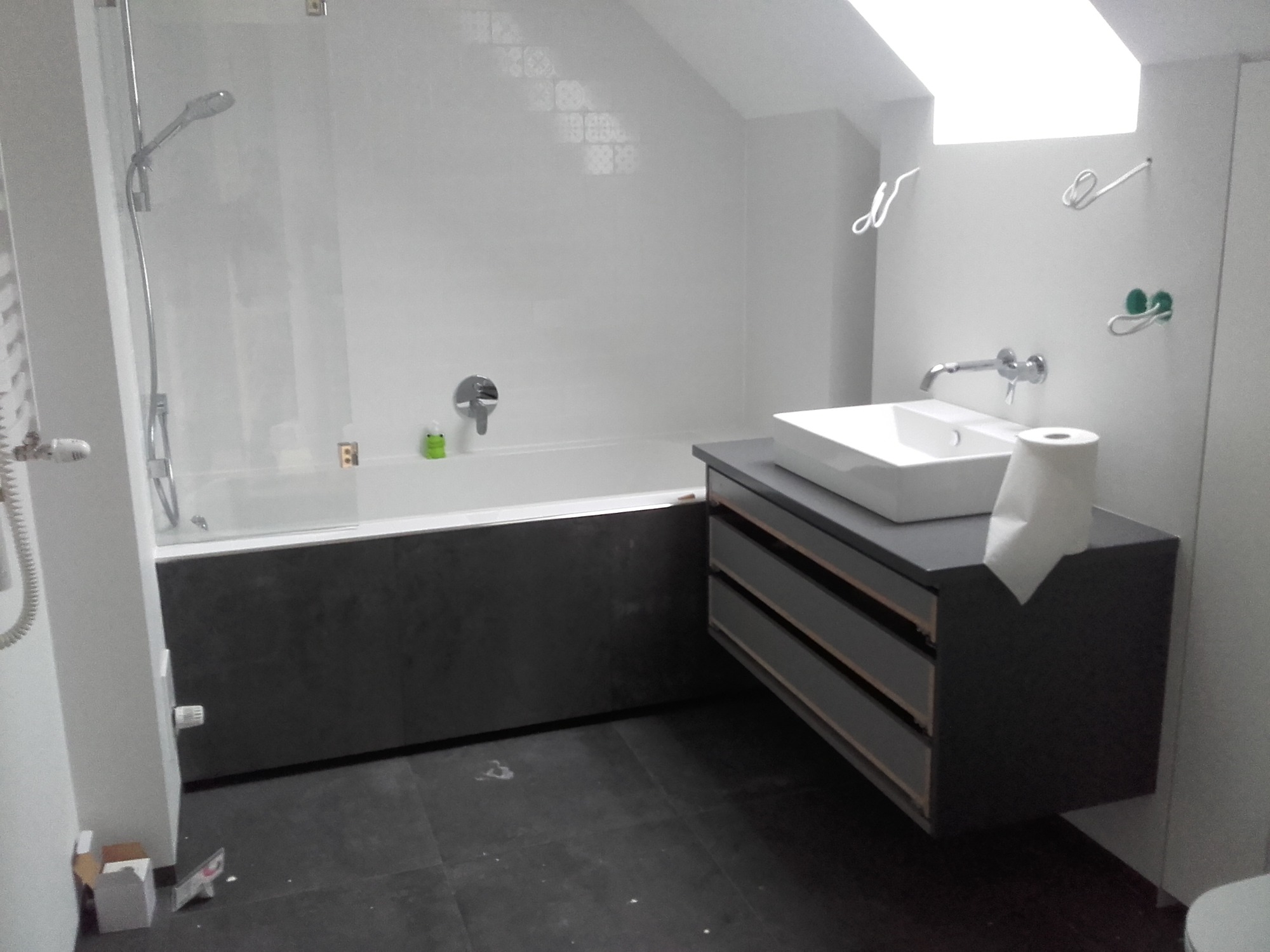 Wykończona łazienka na poddaszu z wanną z szarą obudową, umywalką na szafce z szufladami i szarą podłogą z płytek.