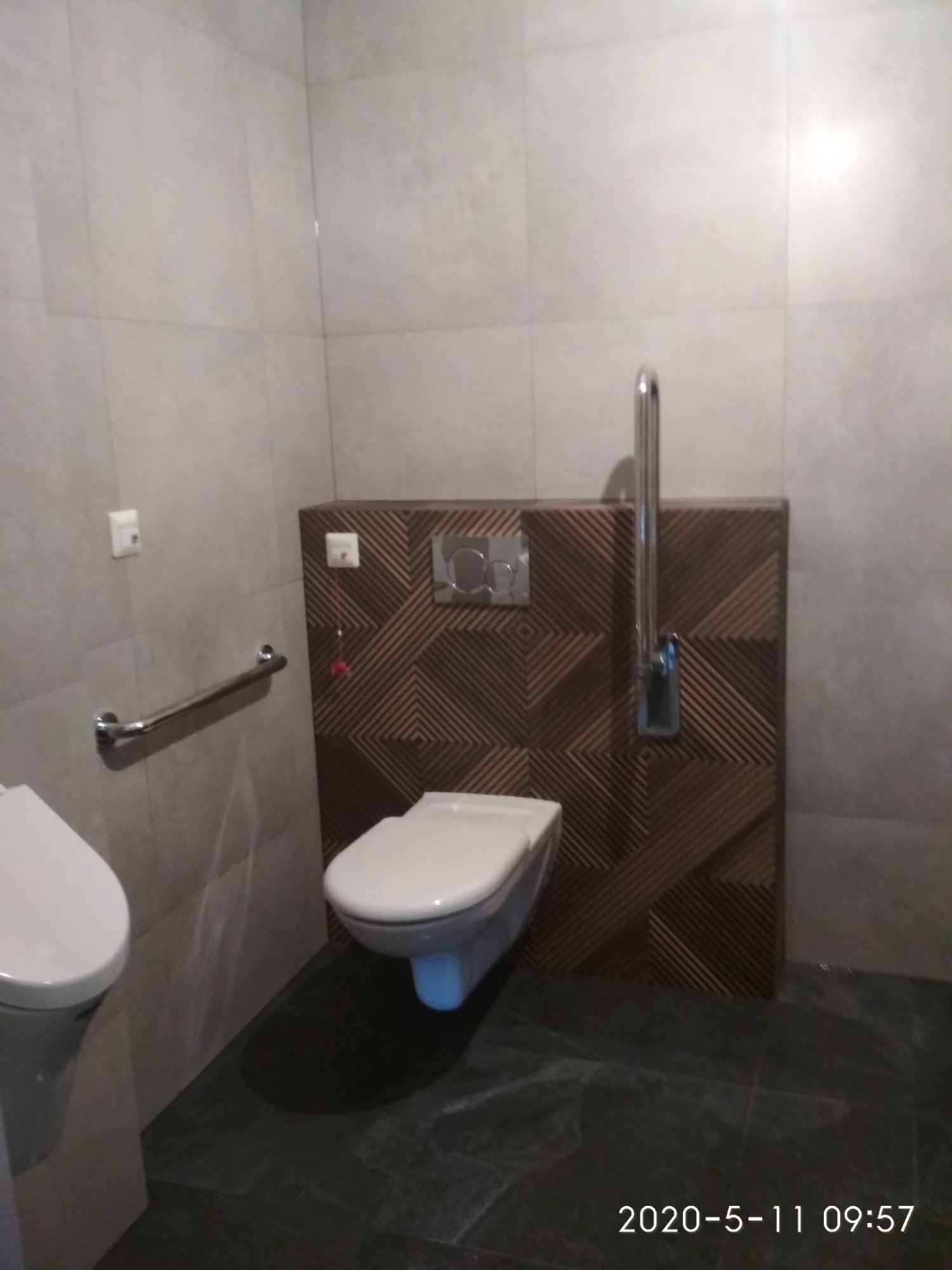 Toaleta dla osób z niepełnosprawnościami z uchwytami, wykończona płytkami w odcieniach szarości i brązu z geometrycznym wzorem, widoczny przycisk spłuczki i awaryjny sznurek.