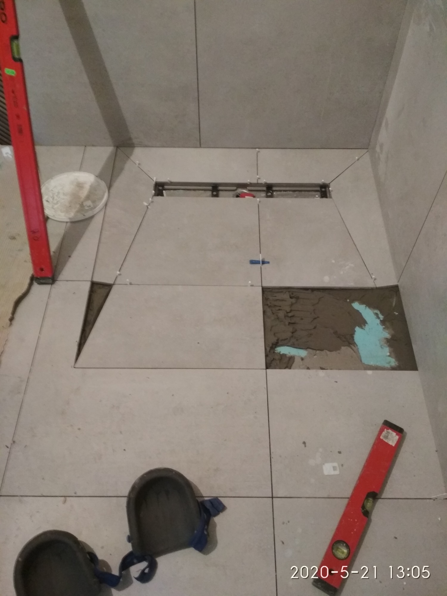 Podczas układania jasnoszarych płytek podłogowych w łazience widoczny jest odpływ liniowy, poziomica oparta o ścianę, klej z niebieskim barwnikiem pod płytkami i nakolanniki glazurnicze.