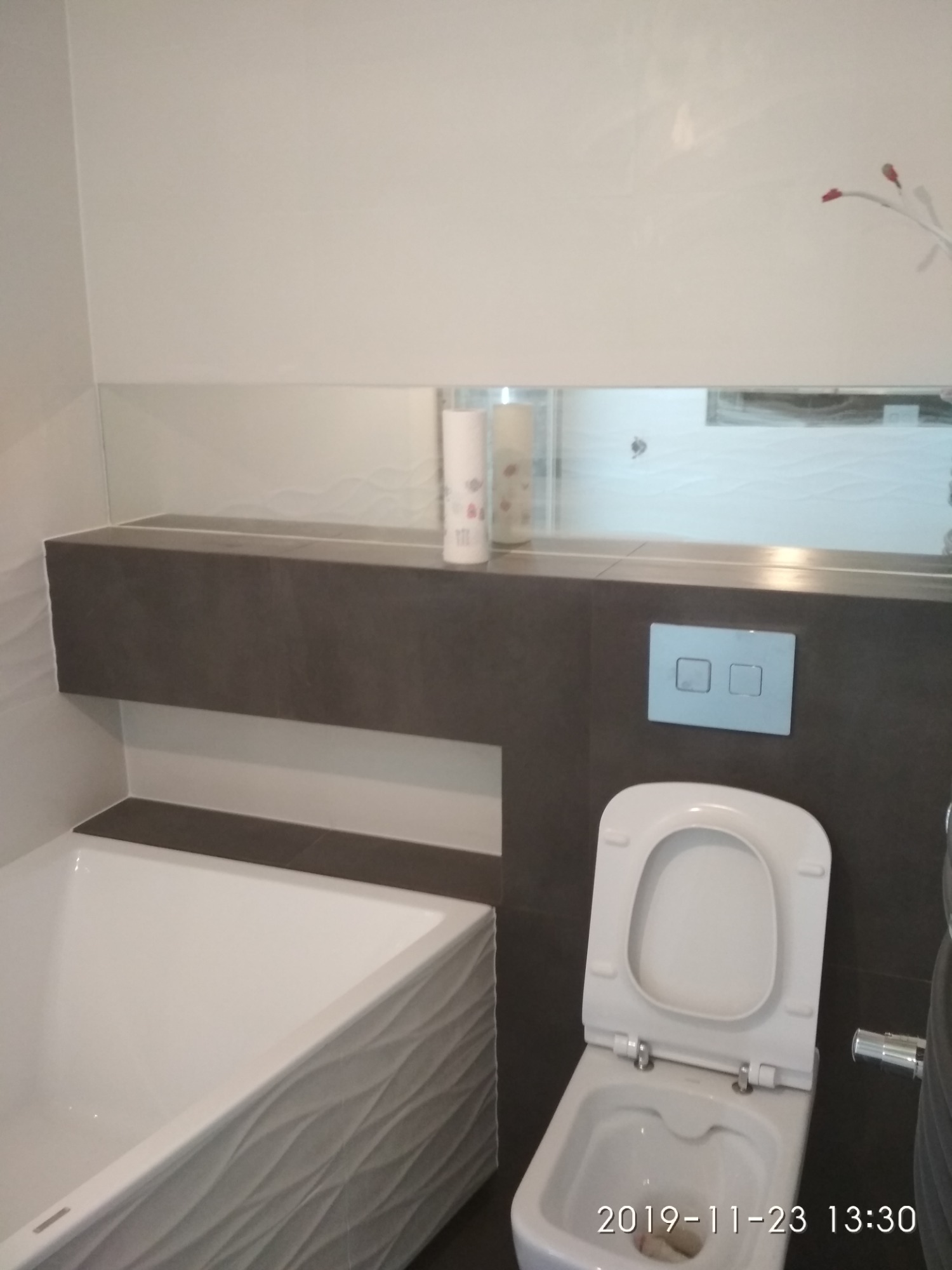 Nowoczesna łazienka z białą wanną o falistej strukturze, podwieszanym WC z podwójnym przyciskiem spłukiwania oraz ciemnoszarą zabudową nad wanną i toaletą z lustrem.