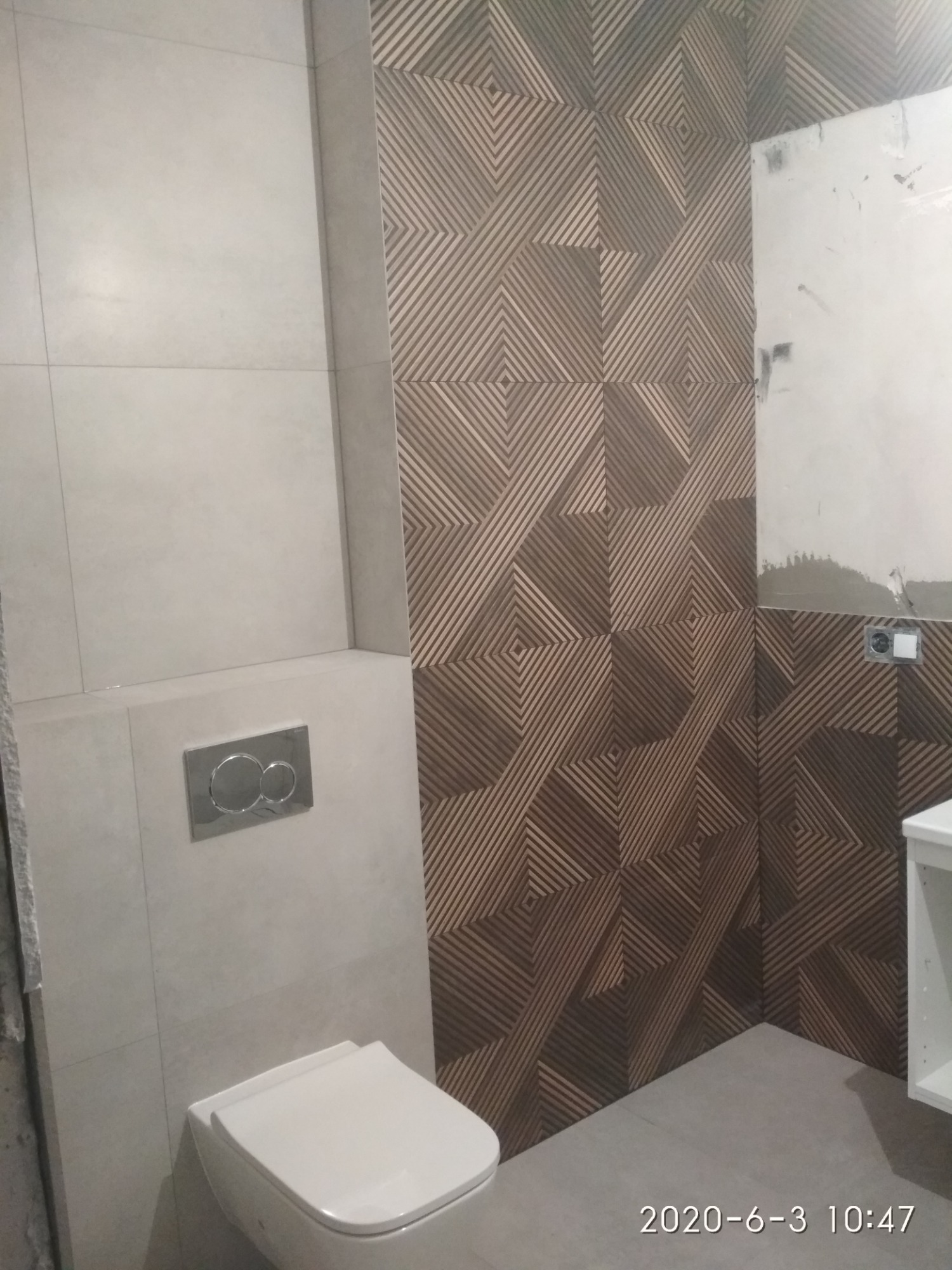 Wykończona łazienka z geometrycznym wzorem płytek na ścianie, widoczny podwieszany sedes i fragment szafki umywalkowej, surowe wykończenie ściany w rogu pomieszczenia.