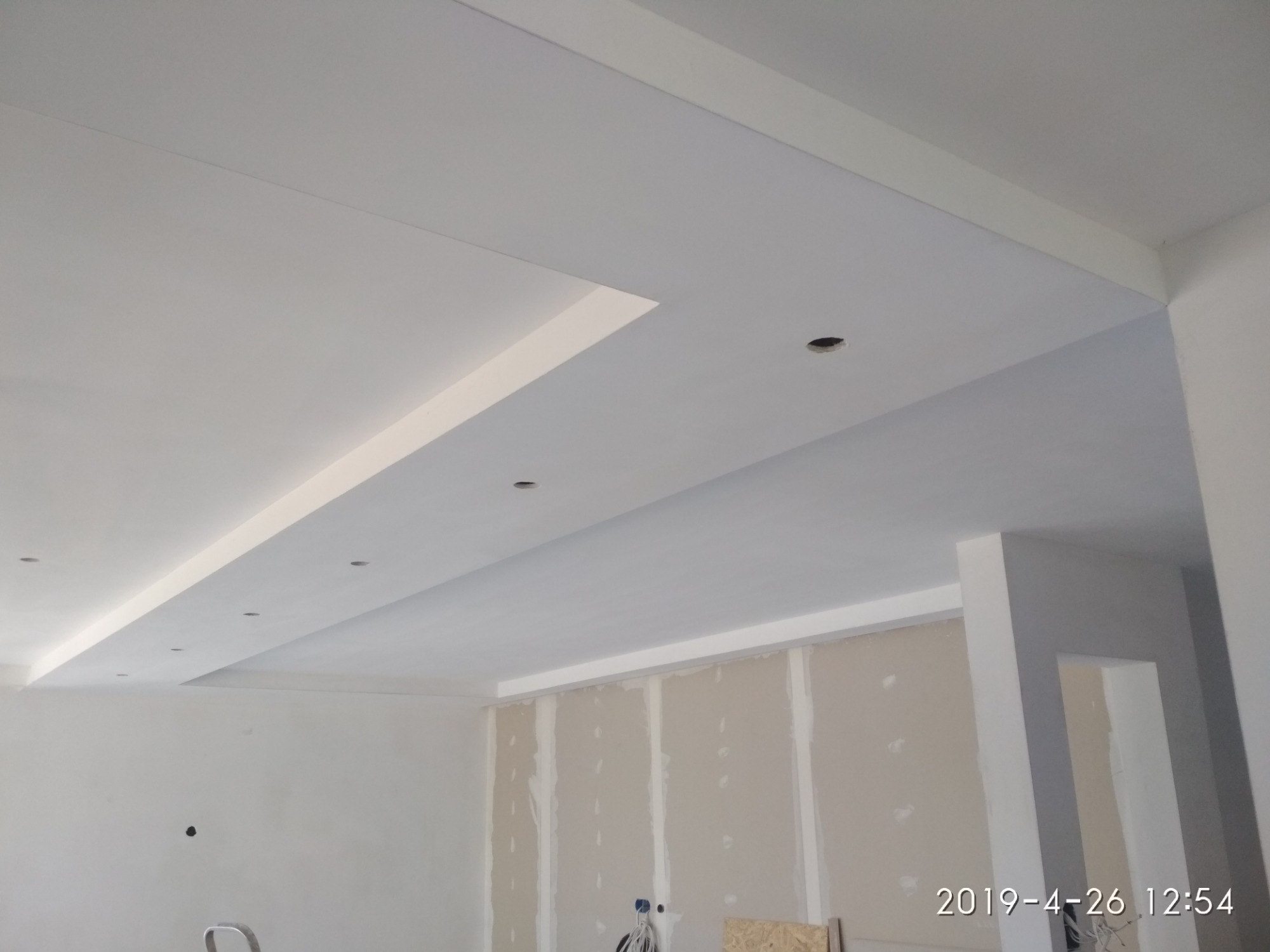 Wnętrze w trakcie prac wykończeniowych: widok na sufit z zabudową z płyt kartonowo-gipsowych i przygotowanymi otworami na oświetlenie punktowe, ściany częściowo pokryte płytami gipsowymi.