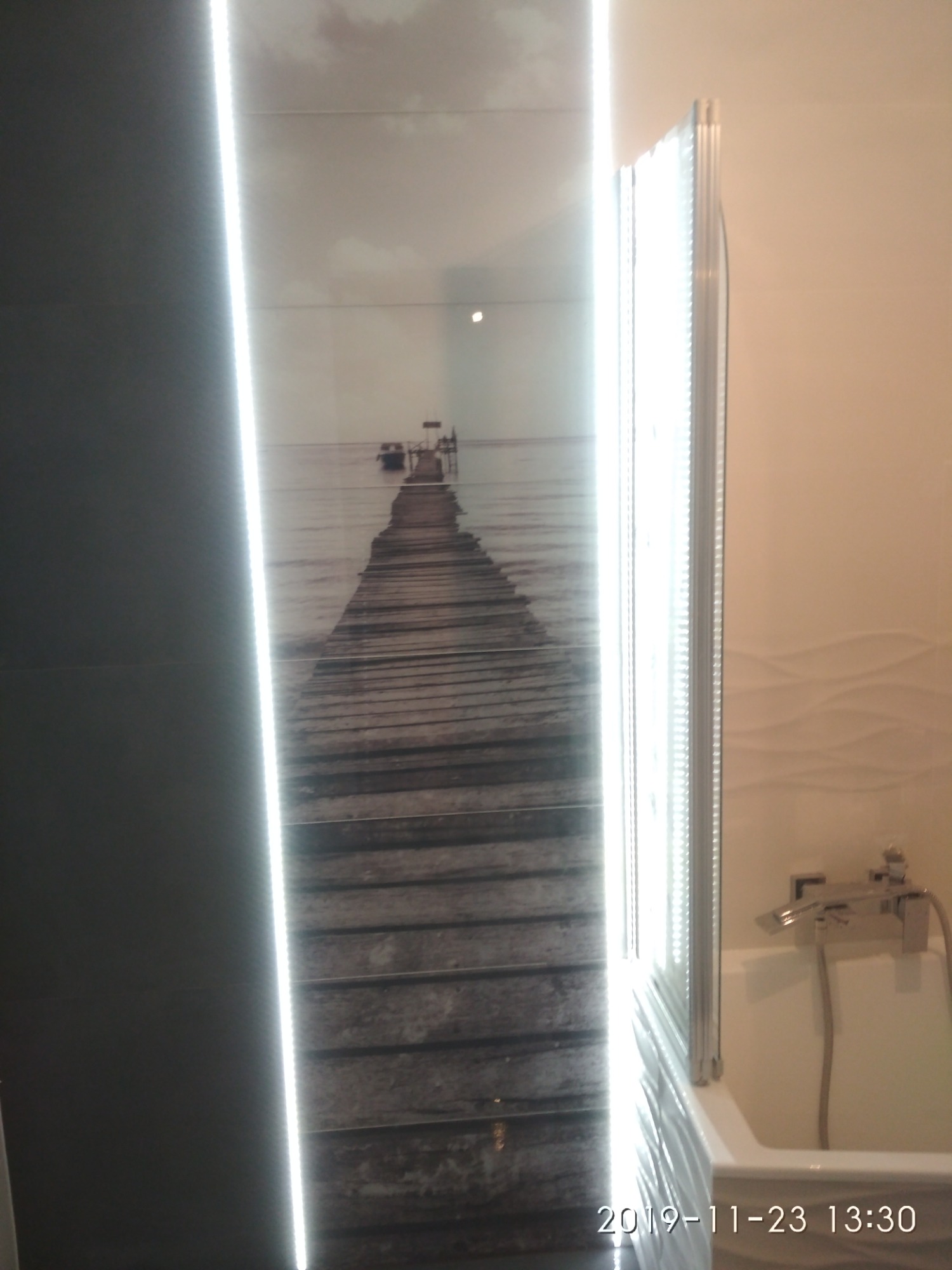 Błyszcząca kabina prysznicowa z widoczną baterią, obok ściana z płytek imitujących drewniany pomost prowadzący do morza, z podświetleniem LED.