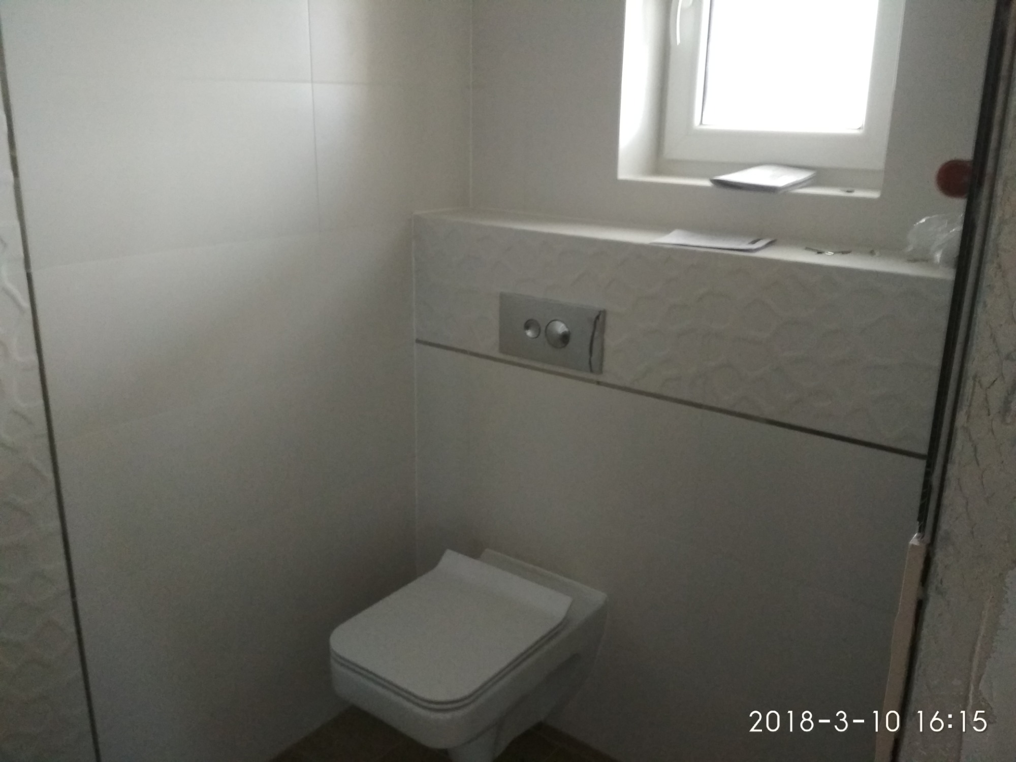 Nowoczesna toaleta podwieszana z białymi płytkami na ścianach i podtynkową spłuczką, widoczna przez częściowo otwarte drzwi.