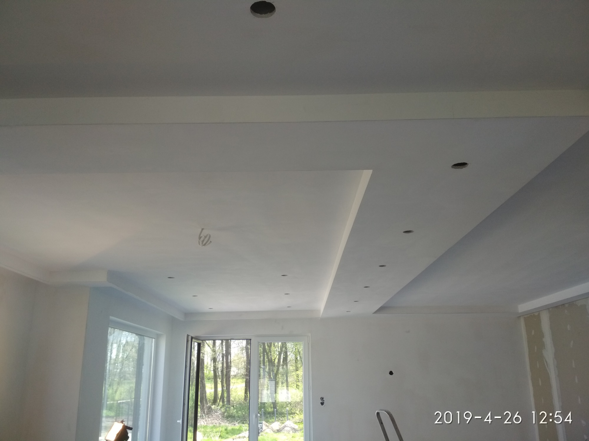 Wykończony sufit podwieszany z otworami na oświetlenie punktowe i ozdobnymi listwami, widok z dołu na jasne wnętrze z oknami wychodzącymi na zieleń.