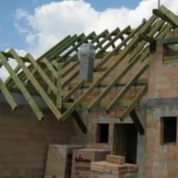 Konstrukcja drewniana dachu w trakcie budowy, widoczne pustaki ceramiczne, komin w szarej izolacji oraz materiały budowlane na paletach.