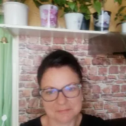 Portret kobiety w okularach na tle półki z doniczkami i tapety imitującej cegłę.