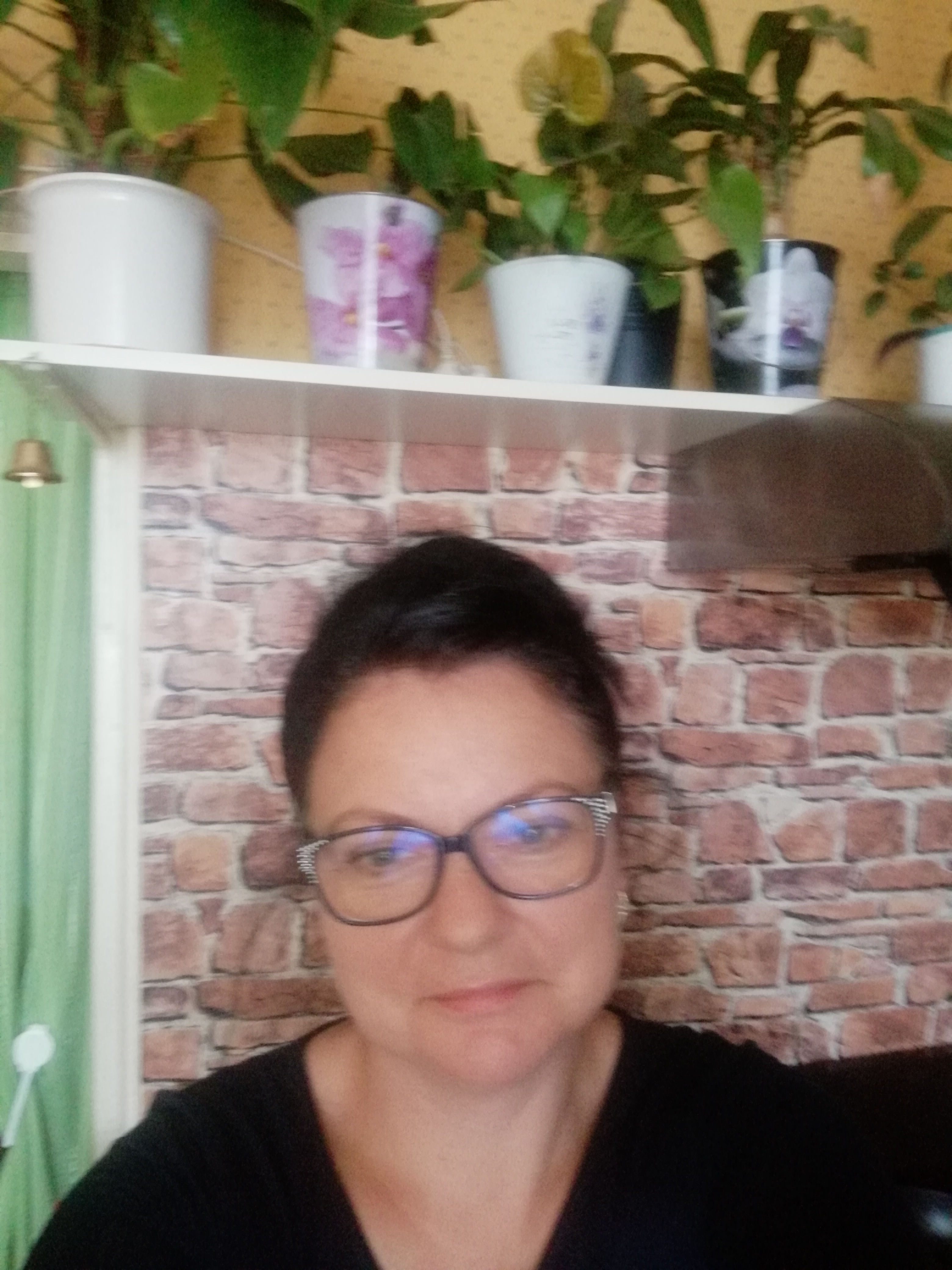 Portret kobiety w okularach na tle półki z doniczkami i tapety imitującej cegłę.