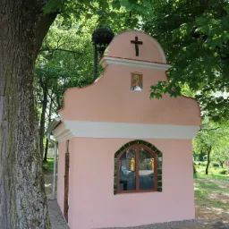Różowa kapliczka z krzyżem i obrazem religijnym, otoczona zielenią drzew, z widocznym fragmentem pnia drzewa z przodu.