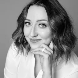 Portret uśmiechniętej kobiety w białej koszuli, z dłonią pod brodą, w czarno-białej tonacji, z widocznym zegarkiem na nadgarstku i białym pierścionkiem.