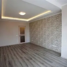 Pusty pokój po remoncie: szare panele podłogowe, ściana z tapetą w ornamenty, podwieszany sufit z oświetleniem LED, białe ściany i drzwi z szybą.