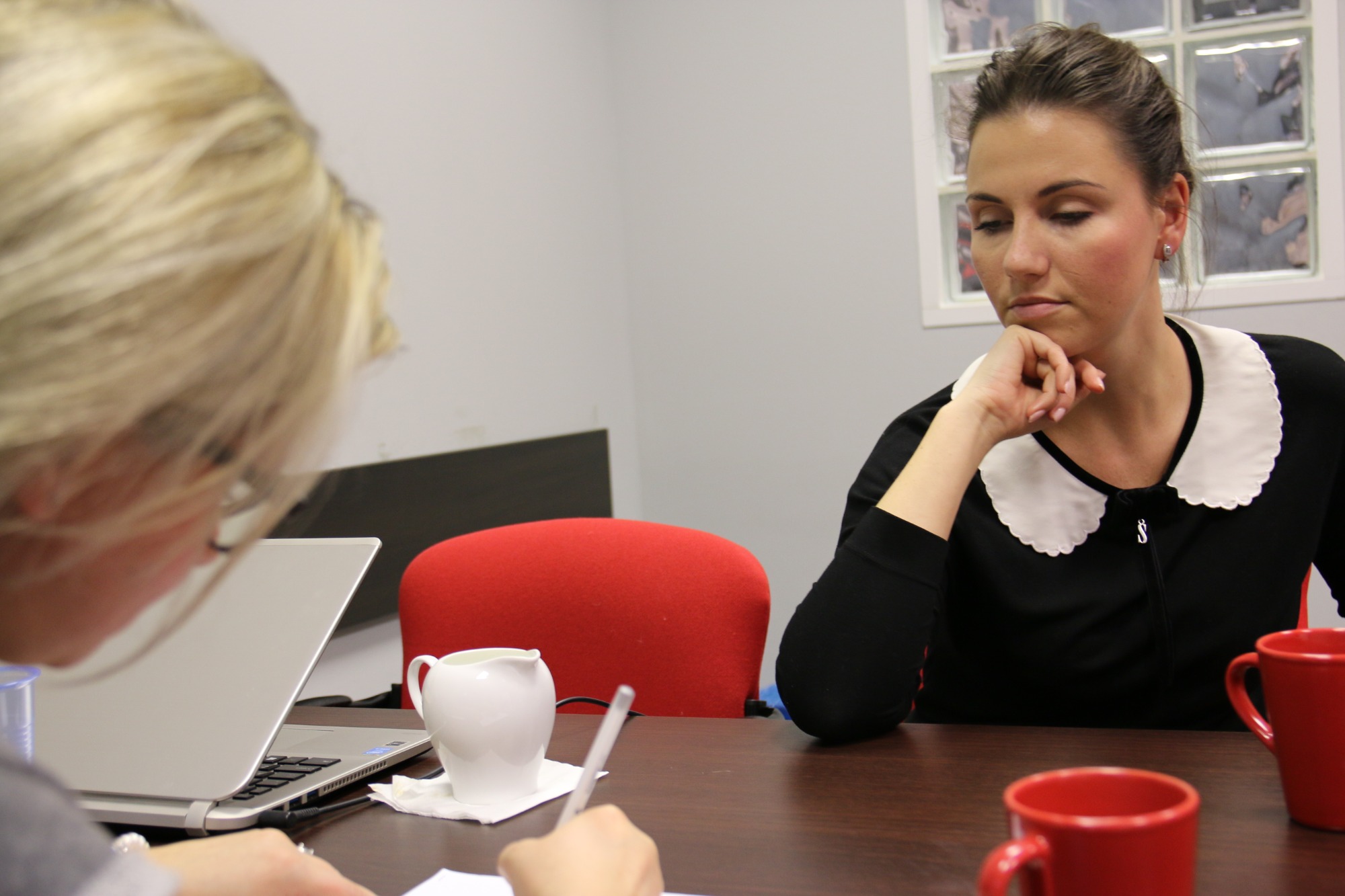 Kobieta w czarnej bluzce z białym kołnierzykiem, zamyślona, siedzi przy biurku podczas spotkania, obok laptop, notatki i czerwone kubki.