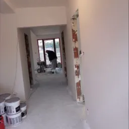 Malowanie ścian w trakcie remontu: widoczny pracownik z agregatem malarskim, puszki z farbą na podłodze, świeżo pomalowane ściany i sufit w jasnym odcieniu oraz surowe ościeżnice drzwiowe.