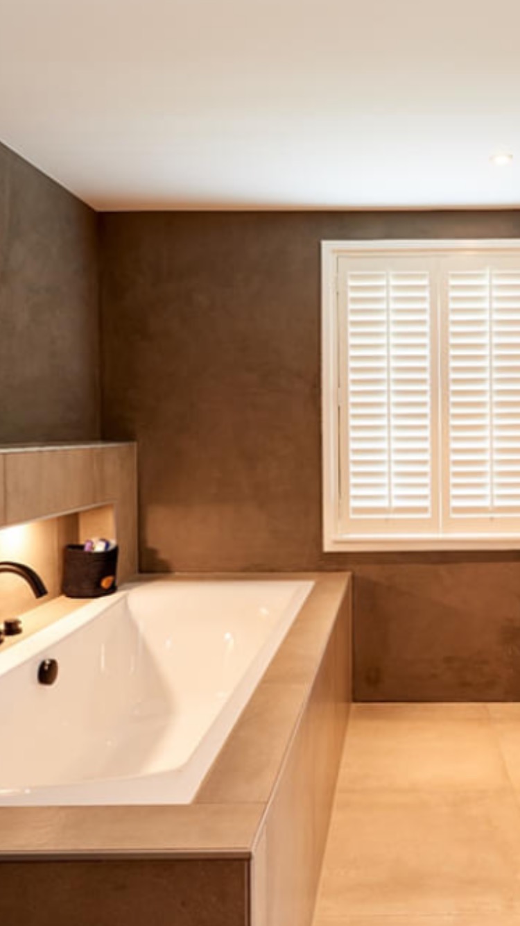 Nowoczesna łazienka z zabudowaną wanną, brązowe ściany z fakturą, białe okno z żaluzjami i beżowa podłoga z płytek.