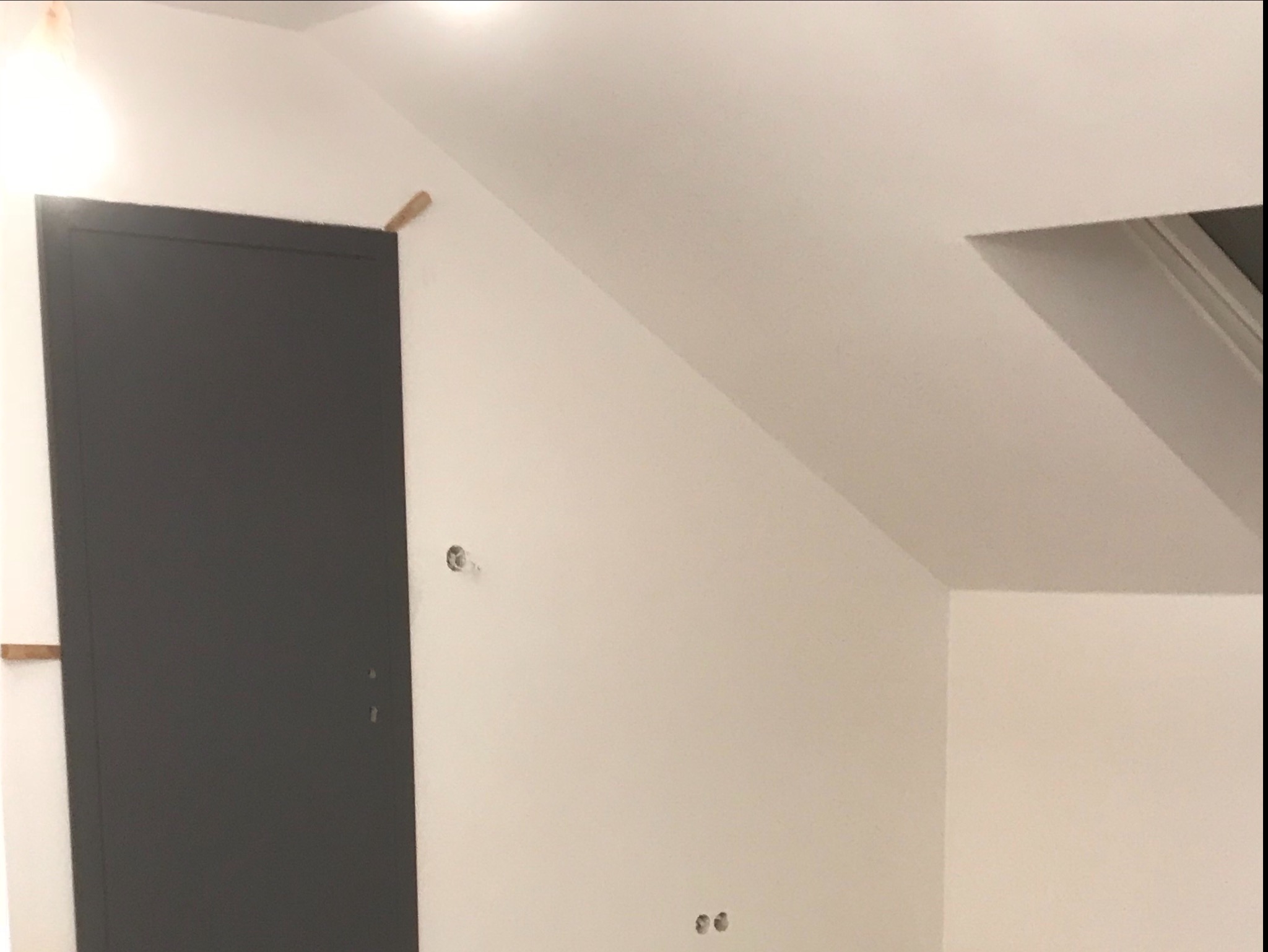 Szara ościeżnica drzwiowa zamontowana w skosie poddasza, widoczne drewniane rozpórki, białe ściany w trakcie wykańczania, otwór okienny na poddaszu.