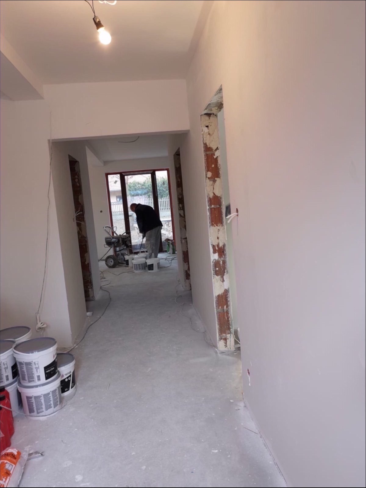 Malowanie ścian w trakcie remontu: widoczny pracownik z agregatem malarskim, puszki z farbą na podłodze, świeżo pomalowane ściany i sufit w jasnym odcieniu oraz surowe ościeżnice drzwiowe.