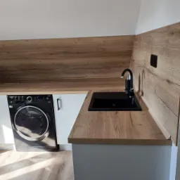 Aranżacja przestrzeni z pralką w zabudowie pod blatem drewnopodobnym z czarnym zlewem i baterią, jasne wnętrze ze skośnym sufitem.