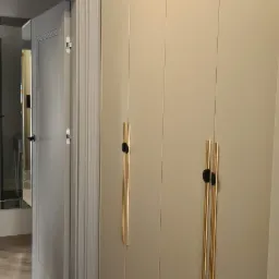 Beżowa szafa wnękowa z złotymi uchwytami na tle marmurowej podłogi i otwartych drzwi do innego pomieszczenia.