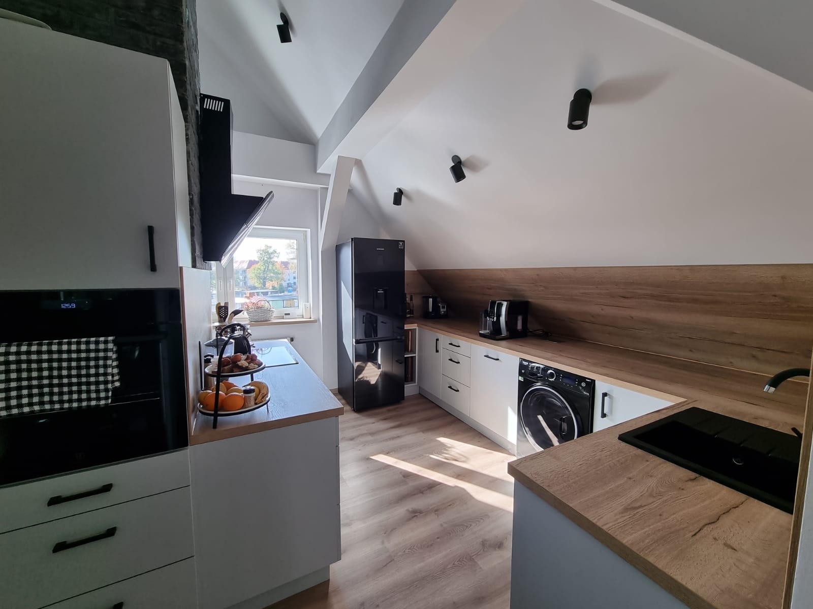 Nowoczesna kuchnia w skosach na poddaszu z białymi szafkami, drewnianym blatem i czarnym sprzętem AGD, w tym pralka w zabudowie.