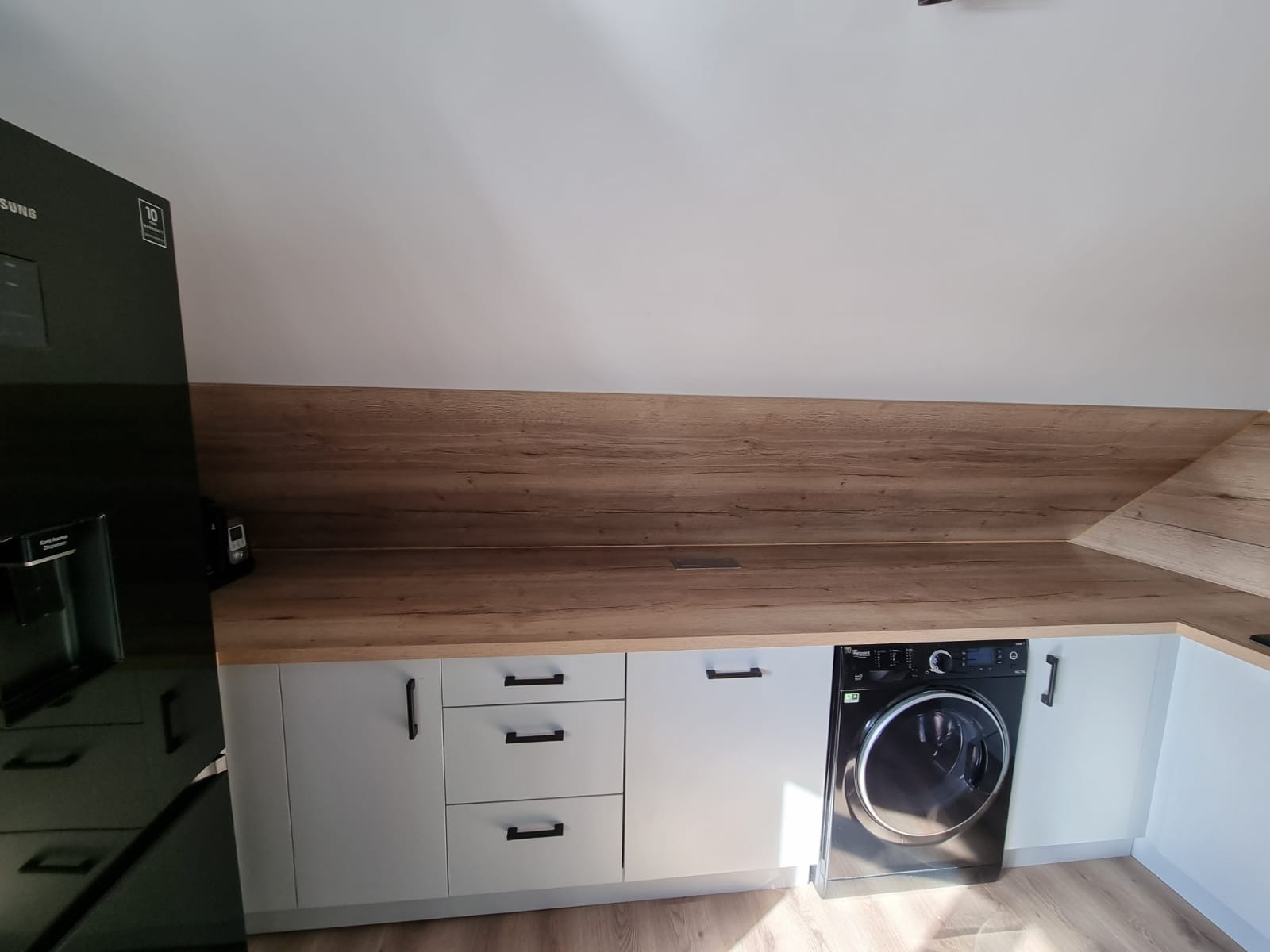 Szare szafki kuchenne z drewnianym blatem w skosie, zintegrowane z czarną pralką automatyczną i czarną lodówką Samsung po lewej stronie.