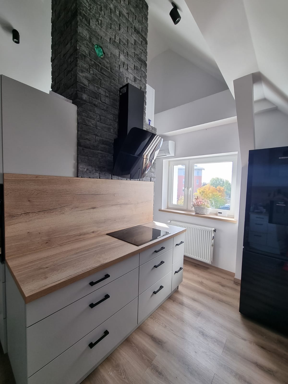 Nowoczesna kuchnia z szarymi szafkami, drewnianym blatem i okładziną ścienną imitującą cegłę, czarny okap, widok na jesienne drzewa za oknem.