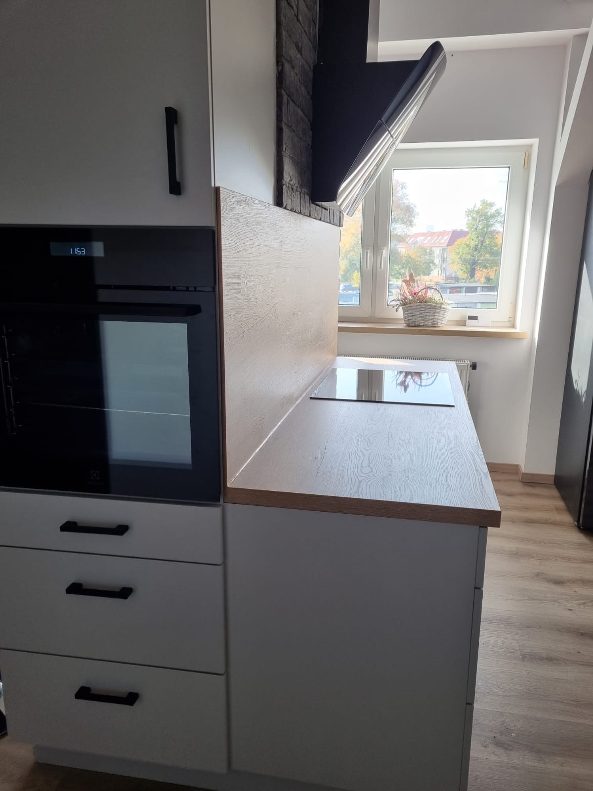 Fragment nowoczesnej kuchni z szarymi szafkami, drewnianym blatem i płytą grzewczą, widok na okno z dekoracyjnym koszykiem.