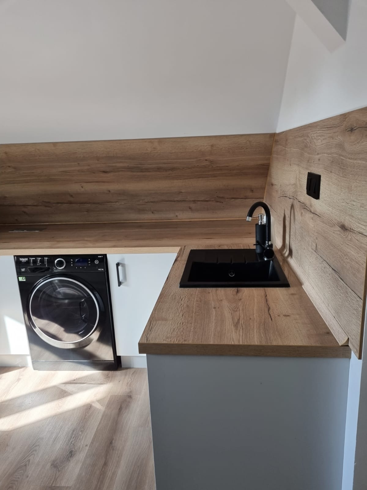 Aranżacja przestrzeni z pralką w zabudowie pod blatem drewnopodobnym z czarnym zlewem i baterią, jasne wnętrze ze skośnym sufitem.
