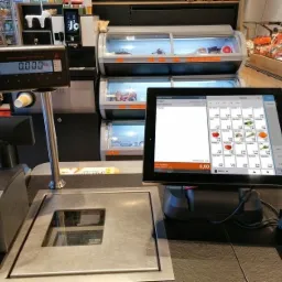 Stanowisko kasowe z wagą, terminalem płatniczym i ekranem dotykowym w sklepie spożywczym, na tle regałów chłodniczych z produktami.